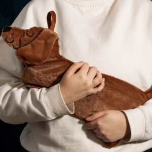 Close-up of a person wearing a white sweater gently hugging a long, brown, plush hot water bottle cover shaped like a smiling Dachshund (sausage dog). Ideal cozy gift for animal lovers or for use during cold weather.