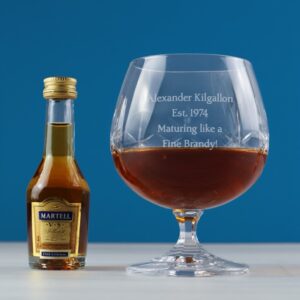 A large, personalised cut crystal brandy snifter glass filled with amber brandy, standing next to a miniature bottle of Martell VS Fine Cognac. The glass is engraved with custom text like 'Maturing like a Fine Brandy!' and a name. Ideal luxury gift for him.