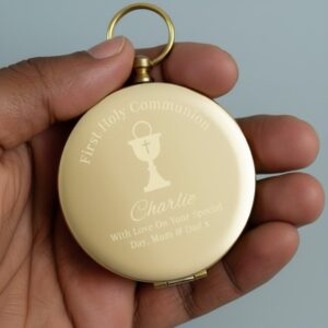 gold-toned personalised compass held in a hand, engraved with 'First Holy Communion' text, a Chalice symbol, the name 'Charlie,' and a loving message 'With Love On Your Special Day, Mum & Dad x'. Ideal sentimental gift for a First Holy Communion.