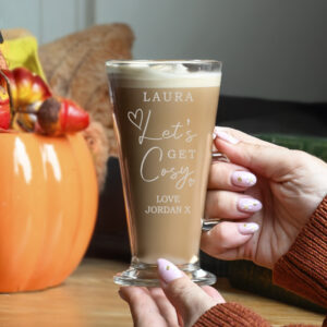 Hand holding a latte glass etched with 'Let's Get Cosy' next to a pumpkin.