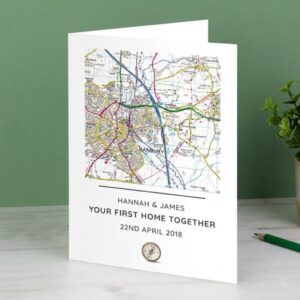 A standing, personalised Ordnance Survey map card on a grey surface, against a green wall, featuring the area around Banbury. Printed text below reads 'HANNAH & JAMES', 'YOUR FIRST HOME TOGETHER', and '22ND APRIL 2018', with a small compass icon at the base.