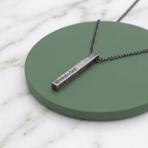 A close-up photograph of a personalised, aged-silver-toned solid bar pendant necklace resting on a circular, matte sage green tray, which is placed on a white marble surface with light grey and gold veining. The rectangular pendant is deeply engraved with the name 'alexander' in a clean font. A matching dark metal box chain is attached and drapes across the tray and marble.