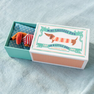A tiny plush dachshund wearing a red and white striped jumper, tucked into a blue polka-dot bed inside a beautifully illustrated matchbox-style house.