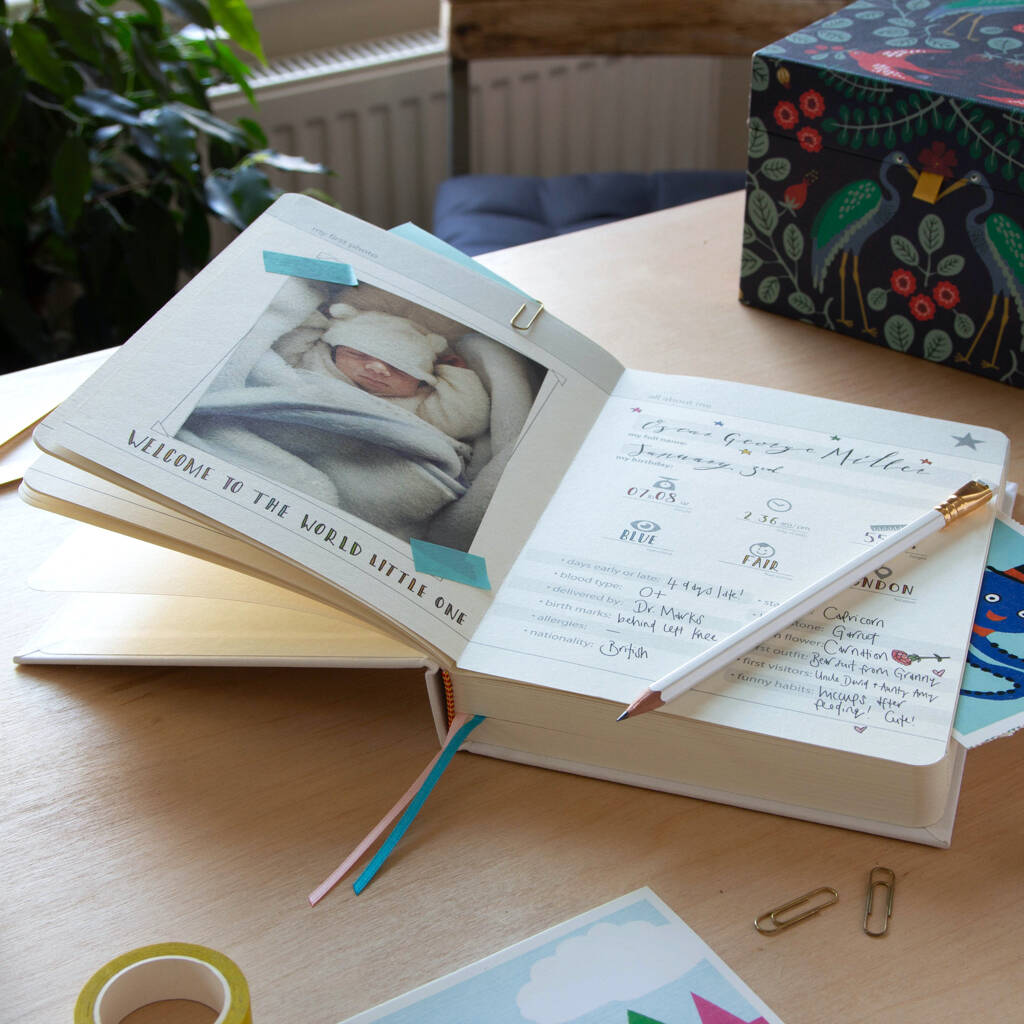 A beautifully designed, open baby journal or memory book, displaying the first page with a photo of a newborn baby wrapped in a blanket. The page includes fill-in sections for the baby's name ('George Miller'), birth details (date, weight, eye colour), and early milestones. Ideal keepsake gift for new parents to record their child's first 26 months.