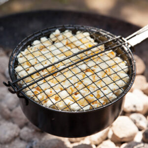 A round, black metal popcorn pan with a long handle and a secure mesh lid, holding freshly popped white popcorn kernels. The pan is held directly over glowing charcoal briquettes in an outdoor barbecue or fire pit. The close-up view emphasizes the rugged cooking tool and the successful, fluffy result. Ideal accessory for camping, grilling, or outdoor entertaining.