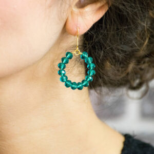 Close-up shot of a woman wearing a pair of gold-hooked hoop earrings. The hoop shape is formed by a cluster of faceted, vibrant emerald green glass beads, creating a colourful statement dangle earring.