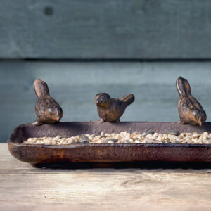 Close-up of a rustic, cast iron bird feeder tray filled with sunflower seeds and nuts, featuring three decorative bird sculptures perched on the rim. The feeder sits on a wooden surface against a blurred blue wooden fence background. Ideal wildlife garden gift.