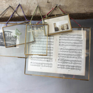 A group of several Brass Hanging Picture Frames of different sizes and orientations (portrait and landscape) suspended from a wooden beam by colourful recycled sari ties. The transparent glass frames display various items, including a vintage postcard, dried white flowers, a botanical print, and antique sheet music, creating a unique, overlapping collage.