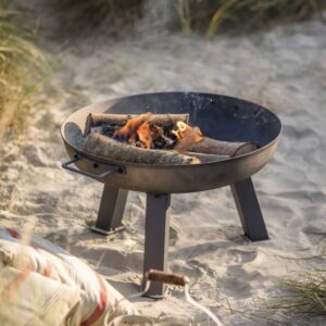 A small, dark grey cast iron fire pit elevated on three sturdy legs, shown burning logs with visible flames, set on a sandy beach or dune with tall grass and a striped picnic blanket visible in the foreground.
