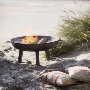 A medium-sized, dark cast iron fire pit sitting on a tripod base, actively burning logs with bright flames, set outdoors on a sandy beach next to tall dune grass and two striped cushions on the sand.