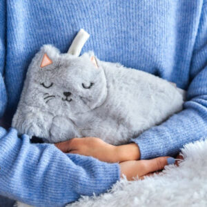 A person wearing a cozy blue jumper is cradling a fluffy, grey hot water bottle cover shaped like a sleeping cat in their hands. The cat cover has embroidered sleepy eyes, whiskers, and small pink ears. The image highlights warmth, comfort, and the soft, plush texture. Ideal gift for cat lovers, Christmas, or a cosy self-care present.