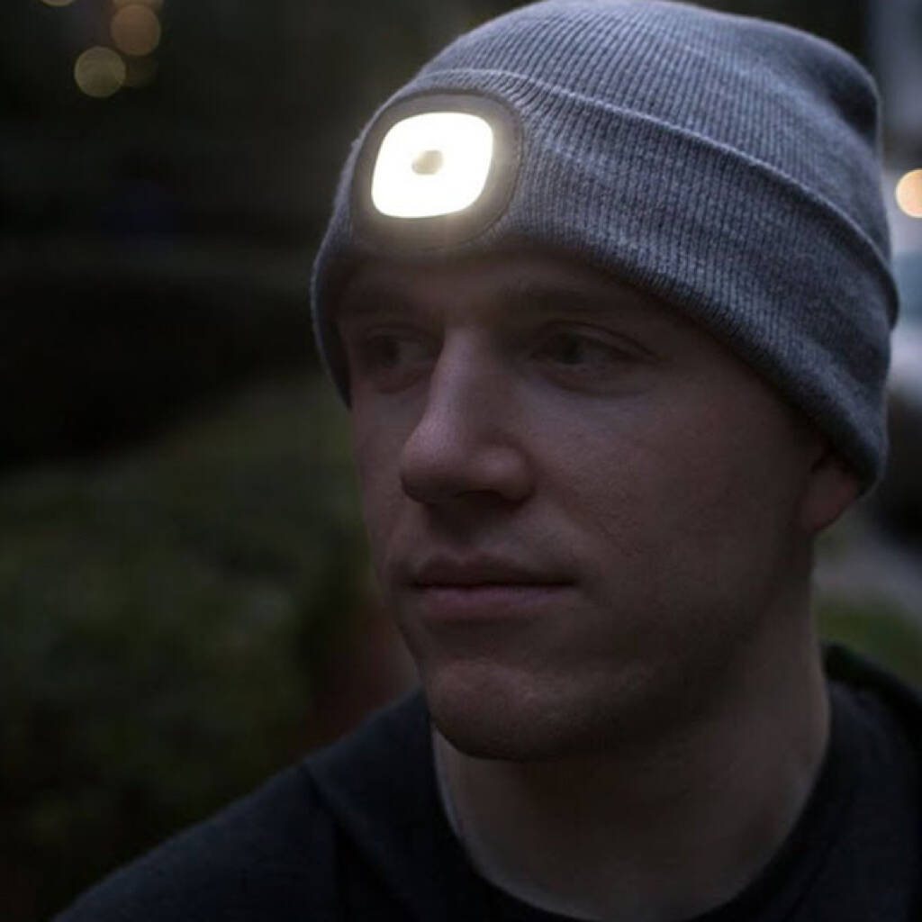 A man wearing a grey knitted beanie hat with a bright, square LED light panel embedded in the forehead, facing forward in a dimly lit, outdoor setting. The light is switched on for visibility. Ideal practical gift for cyclists, runners, hikers, or anyone needing hands-free light in winter.