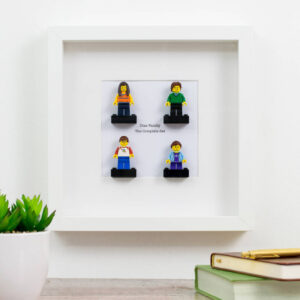 A white-framed display piece featuring four custom mini figures standing on black platforms. Below the figures, the text "Diaz Family The Complete Set" is printed, suggesting a family or group portrait made from personalized toy bricks. The frame is shown sitting on a desk next to a green potted plant and books. Ideal personalised family gift, Mother's Day, Father's Day, or anniversary present for fans of brick toys.