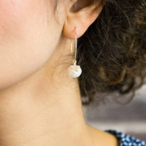 Close-up shot of a woman wearing a single silver hoop earring with a dangling, pale iridescent freshwater pearl charm. The pearl is topped with a small, gold-tone star detail. The earring is being worn on the right ear, framed by dark, curly hair.