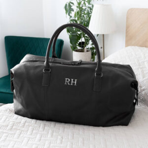 A black vegan leather weekender holdall featuring the initials "RH" monogrammed in silver on the front, placed on a white bedspread with a lamp and green plant in the background.