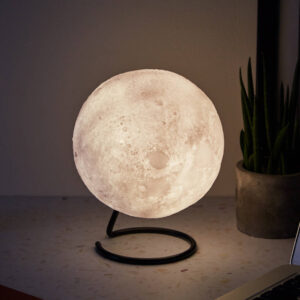 A realistic, detailed 3D orb moon lamp glowing softly on a simple black metal spiral stand, sitting on a table next to a green plant in a concrete pot.