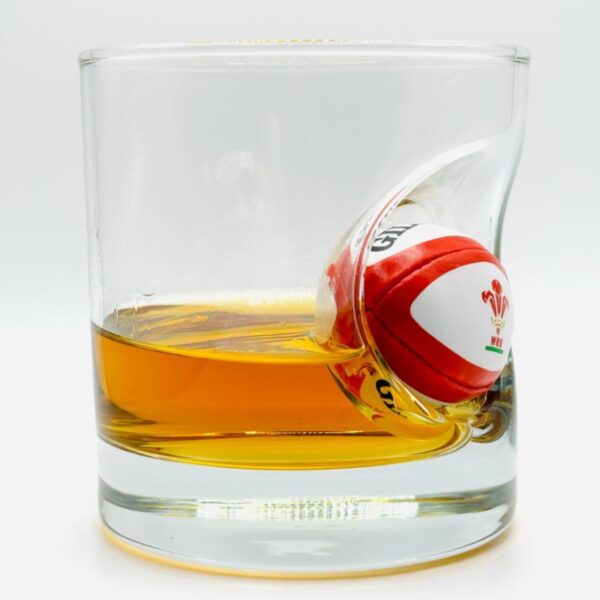 Heavy whisky tumbler filled with amber liquor featuring an embedded miniature red and white Wales WRU rugby ball.