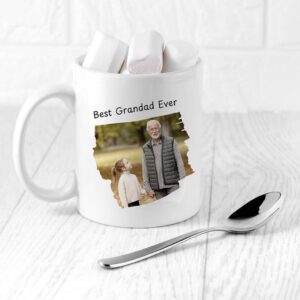 A white ceramic mug personalised with the text "Best Grandad Ever" printed above a photo of a smiling grandad holding hands with his granddaughter in an outdoor, autumnal setting. The mug is sitting on a white wooden surface with a chrome spoon beside it, and is filled with three large marshmallows.