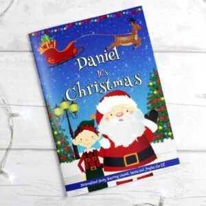 Personalised Boys 'It's Christmas' story book cover featuring the name 'Daniel', Santa Claus, an elf, a flying reindeer and sleigh, set against a snowy night sky, lying on a white wooden surface with fairy lights.
