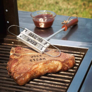 A custom branding iron kit used to sear the name 'Joe's Steak' onto a sizzling T-bone steak cooking on a gas grill. The metal letter tray is suspended over the meat, with barbecue sauce visible in the background. Ideal personalised gift for a grill master, chef, or dad who loves BBQ.
