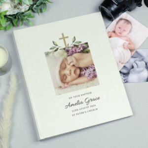 A cream-coloured, personalised photo album for a Christening or Baptism. The cover features a gold cross design with green foliage framing a square photo of a sleeping baby (Amelia Grace) surrounded by purple flowers. Below the photo, the text reads: "ON YOUR BAPTISM," "Amelia Grace," the date "22ND AUGUST 2024," and "ST PETER'S CHURCH." The album is displayed on a light gray surface next to a candle, greenery, a camera lens, and two loose baby photos.