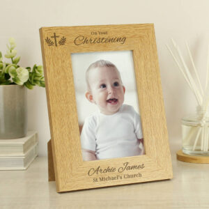 A wooden, landscape-oriented personalised photo frame for a Christening. The oak-effect wood is engraved at the top with a cross and foliage motif above the words "On Your Christening." Below the photo insert of a smiling baby (Archie James), the text is personalised with "Archie James" and "St Michael's Church." The frame is displayed on a light wooden surface next to a plant and a reed diffuser.