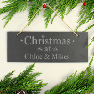 Rectangular personalised hanging slate plaque engraved with 'Christmas at Chloe & Mikes' and a snowflake motif, suspended by twine, surrounded by fresh evergreen garland and red berries.