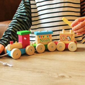 A child wearing a striped shirt plays with a brightly coloured wooden toy train set on a light wooden table. The train carriages are engraved with the name 'Arthur' and a message 'Happy Birthday Love From Mummy & Daddy'. Ideal personalised baby or toddler gift.