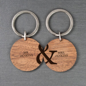 Two round wooden keyrings placed side-by-side on a dark slate background. The left keyring is engraved with MR. JENKINS and the left half of an ampersand. The right keyring is engraved with MRS. JENKINS and the right half of an ampersand, creating a complete design when joined.
