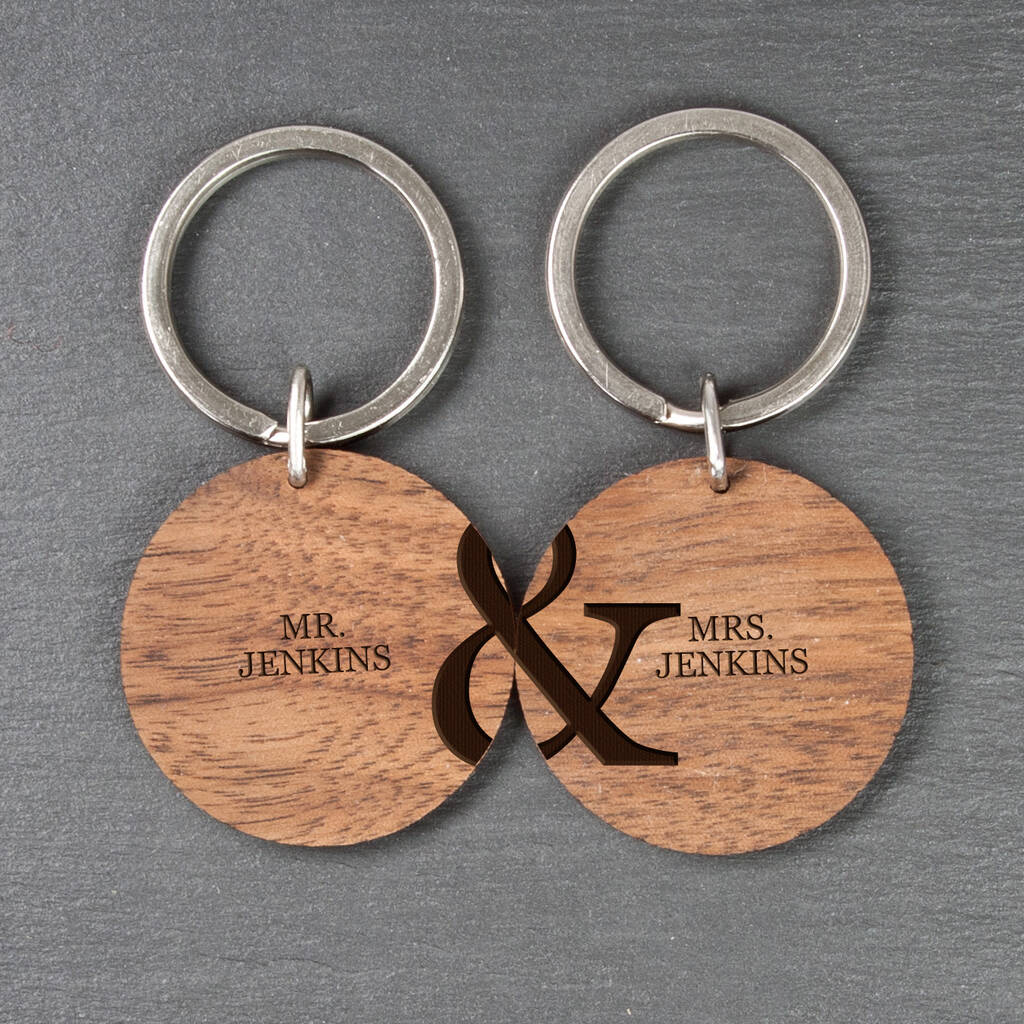 Two round wooden keyrings placed side-by-side on a dark slate background. The left keyring is engraved with MR. JENKINS and the left half of an ampersand. The right keyring is engraved with MRS. JENKINS and the right half of an ampersand, creating a complete design when joined.