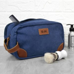 A blue denim wash bag with leather detailing and handles, featuring the initials 'D.H' on a leather patch. A black comb protrudes from the top of the bag, and a white shaving brush sits on the surface next to the bag.