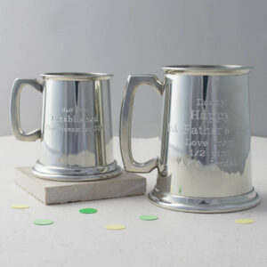 Two highly polished, silver-toned pewter tankards—one half-pint and one full-pint—engraved with personalised messages like 'Happy 1st Father's Day' and 'Established 21st November 2016'. The tankards are displayed on a white surface with confetti. Ideal traditional, engraved gift for Father's Day, Best Man, or Anniversary.