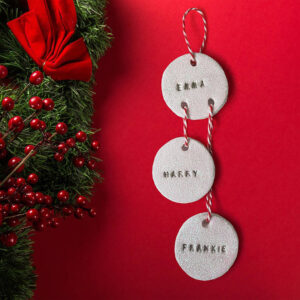 Personalised round family Christmas decoration, made of glittery white clay discs connected by red and white twine, hung against a red background with a festive wreath.
