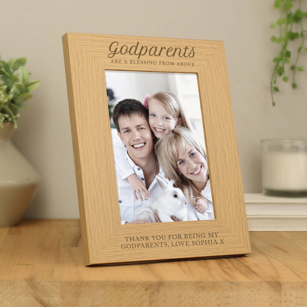 Personalised wooden photo frame engraved with the fixed text "Godparents are a blessing from above" at the top and custom personalised text below the photo, currently showing "Thank You For Being My Godparents, Love Sophia X". The frame holds a 5x7 portrait photo of a smiling man and woman holding a small child and a white rabbit.