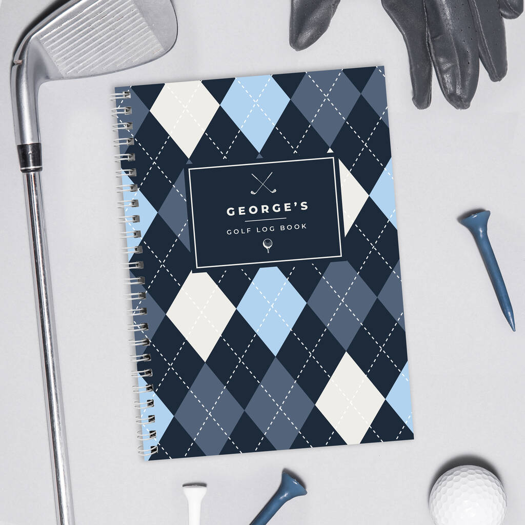 Personalised Golf Log Book featuring a dark blue, light blue, and white argyle pattern cover with a custom name ('George's Golf Log Book'), spiral-bound, surrounded by golf tees, a ball, a glove, and a club head on a grey surface.