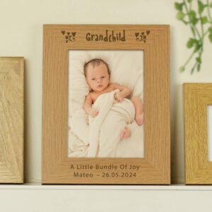 Personalised wooden photo frame with the fixed engraved word "Grandchild" decorated with small hearts and scrolls at the top. The bottom is personalised with the text "A Little Bundle of Joy Mateo - 26.05.2024". The frame holds a 5x7 portrait photo of a newborn baby wrapped in a cream blanket.