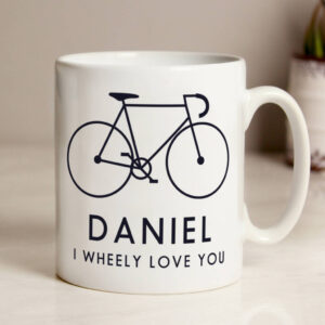 White ceramic mug featuring a black line drawing of a bicycle. Below the bike, the personalised name DANIEL is printed in bold capitals, followed by the pun text I WHEELY LOVE YOU.