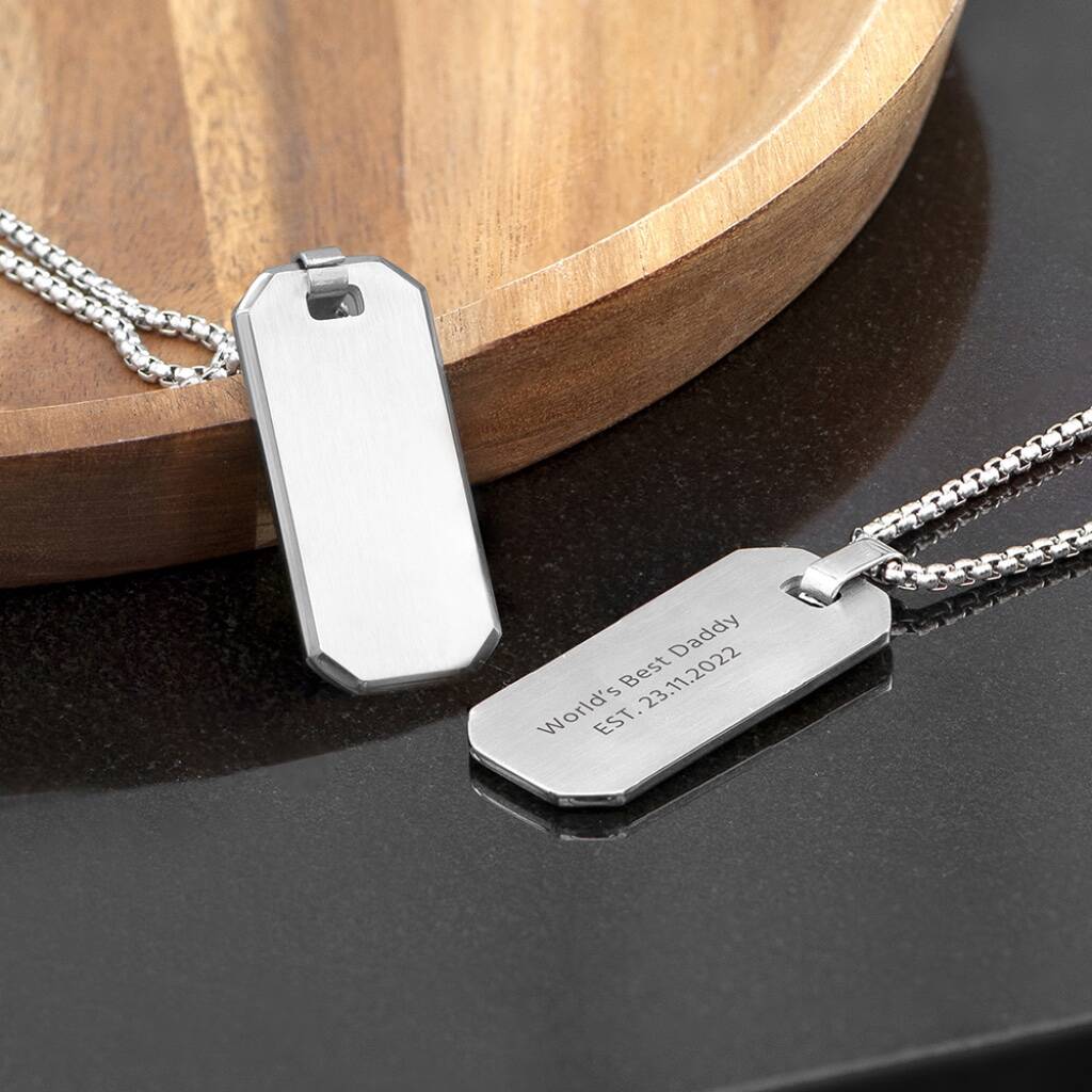 A silver-tone dog tag necklace resting on a polished black surface next to a wooden bowl. The tag is engraved with the text "World's Best Daddy EST. 23.11.2022".