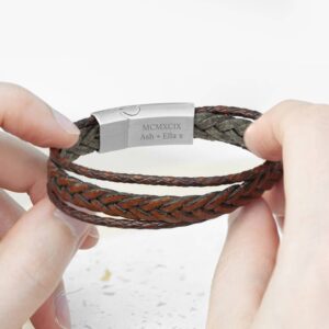 A close-up of a brown layered woven leather bracelet being held, showing the silver stainless steel clasp engraved with 'MCMXCIX Ash + Ella x'.