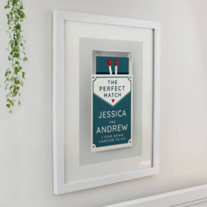White framed wall art featuring a retro teal matchbox illustration with two red matches. Text reads THE PERFECT MATCH, followed by personalised names JESSICA AND ANDREW and a custom message 1 YEAR DOWN FOREVER TO GO.