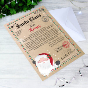 A personalised letter from Santa Claus on thick, vintage-style paper with festive borders, an image of Santa, and the recipient's name ("Thomas") clearly visible, laid diagonally on a wooden surface next to a white envelope and surrounded by subtle fairy lights and iridescent ribbon.