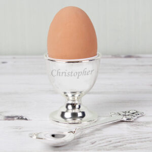 A personalised, polished silver-plated egg cup holding a whole brown egg, sitting on a pale wooden surface next to a matching silver spoon. The egg cup is engraved with the name 'Christopher' in a script font. Ideal traditional and keepsake gift for a child's Christening, first birthday, or Baptism.