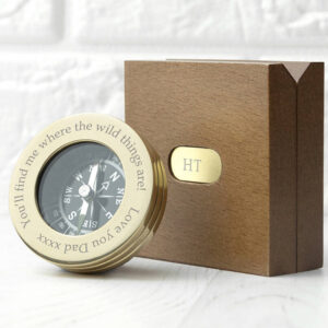 A personalised brass pocket compass resting next to its matching square, brown wooden presentation box, which features a monogrammed brass plate with the initials 'HT'. The compass bezel is engraved with the custom message 'You'll find me where the wild things are! Love You Dad xxxx'. This is an ideal luxury sentimental gift for a traveller, retirement, or graduation.