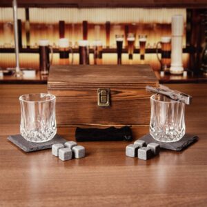 A luxury whisky gift set displayed on a dark wooden bar, featuring a rustic wooden presentation box with a clasp. The set includes two personalised cut crystal-style whisky tumblers resting on black slate coasters, several grey chilling stones, and metal tongs. Ideal engraved gift box for a man, Father's Day, or groomsmen.