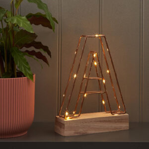 Copper wire frame light up letter 'A' standing on a rectangular light wood base. The letter is illuminated by warm fairy lights inside the frame, sitting next to a potted houseplant with a terracotta pot. Ideal for modern home decor, personalization, or initial display.