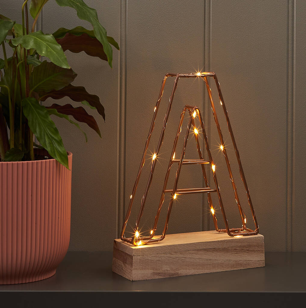 Copper wire frame light up letter 'A' standing on a rectangular light wood base. The letter is illuminated by warm fairy lights inside the frame, sitting next to a potted houseplant with a terracotta pot. Ideal for modern home decor, personalization, or initial display.