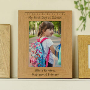 A light oak-coloured wooden photo frame holding a picture of a smiling young girl (like Olivia Ramirez) on her first day of school, wearing a colourful backpack. The frame is engraved with "My First Day at School" at the top, a ruler design, and a personalised name (e.g., Olivia Ramirez) and school name (e.g., Maplewood Primary) at the bottom.
