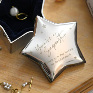 A nickel-plated, star-shaped trinket box with a reflective surface, engraved with the personalised message "You're a Superstar" and "Thank you for being my Bridesmaid," open to show a dark blue velvet interior holding a pearl ring and an earring.