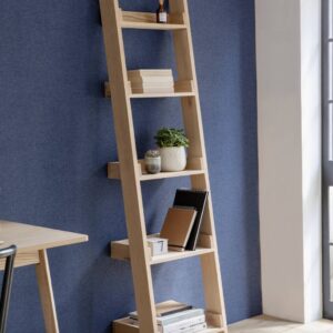 Leaning shelf ladder made from raw light oak with four shelves, holding books, notebooks, and potted plants, set against a dark blue textured wall in a modern home office or living space.