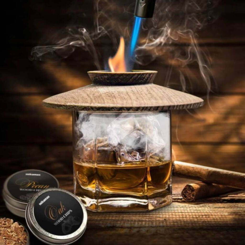 A luxury whisky smoker kit in use: a wooden smoker chimney rests on an ice-filled whisky glass, being ignited by a butane torch to generate wood-chip smoke. Two tins of 'Pecan' and 'Oak' wood chips and a cigar rest nearby on a rustic wooden surface. Ideal gift for whisky connoisseurs.